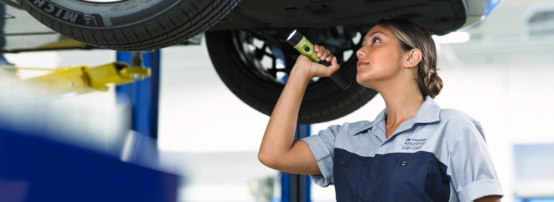 Mechanic inspecting a vehicle's brake system under a lift, using a flashlight to examine the tire and undercarriage.
