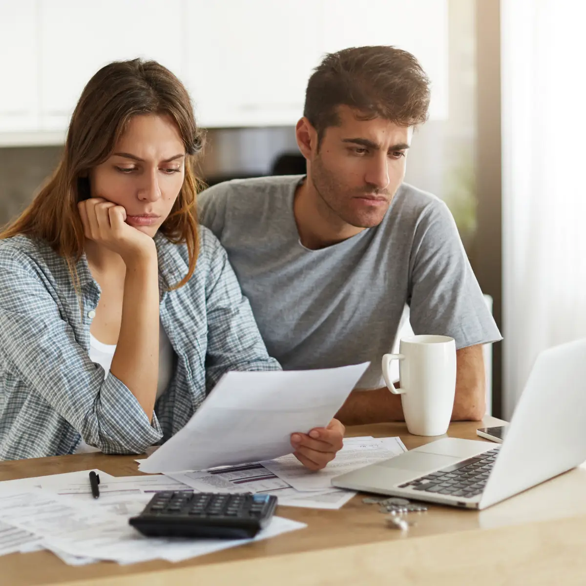 A concerned couple sitting at a table, looking at documents, a laptop, and calculator.