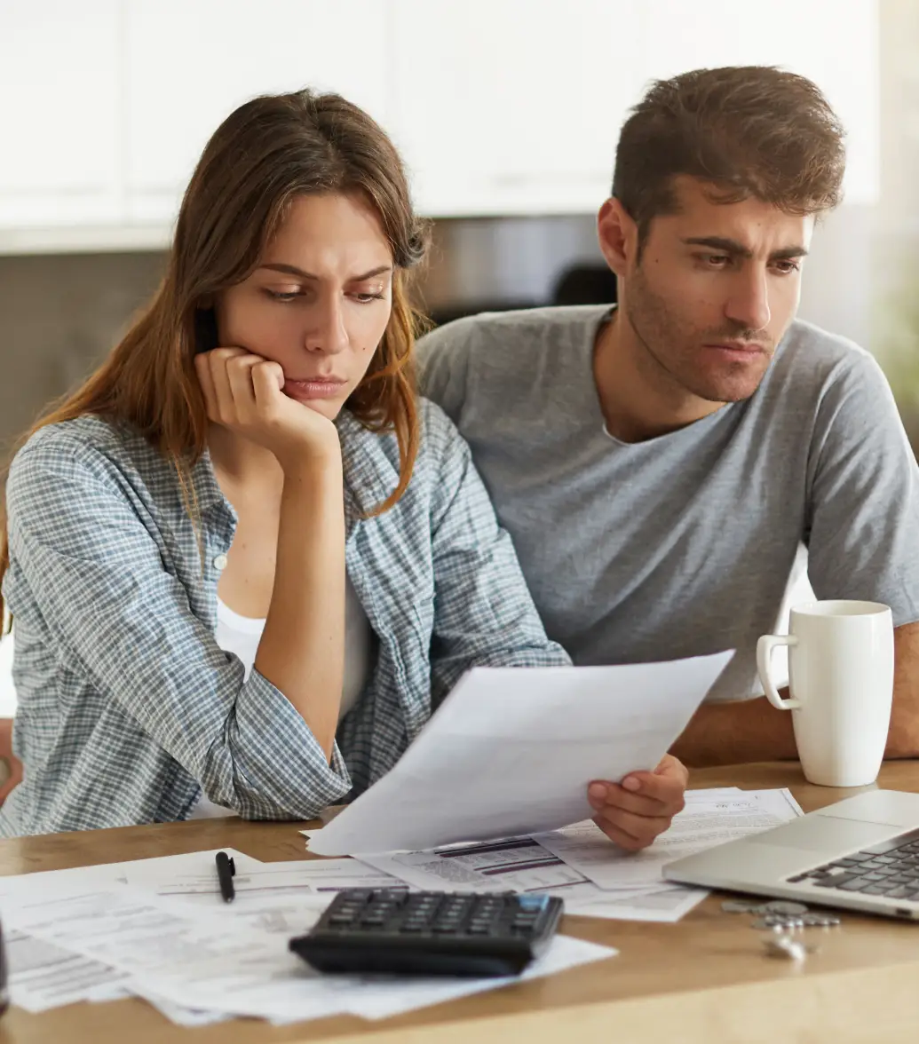 A concerned couple sitting at a table, looking at documents, a laptop, and calculator.