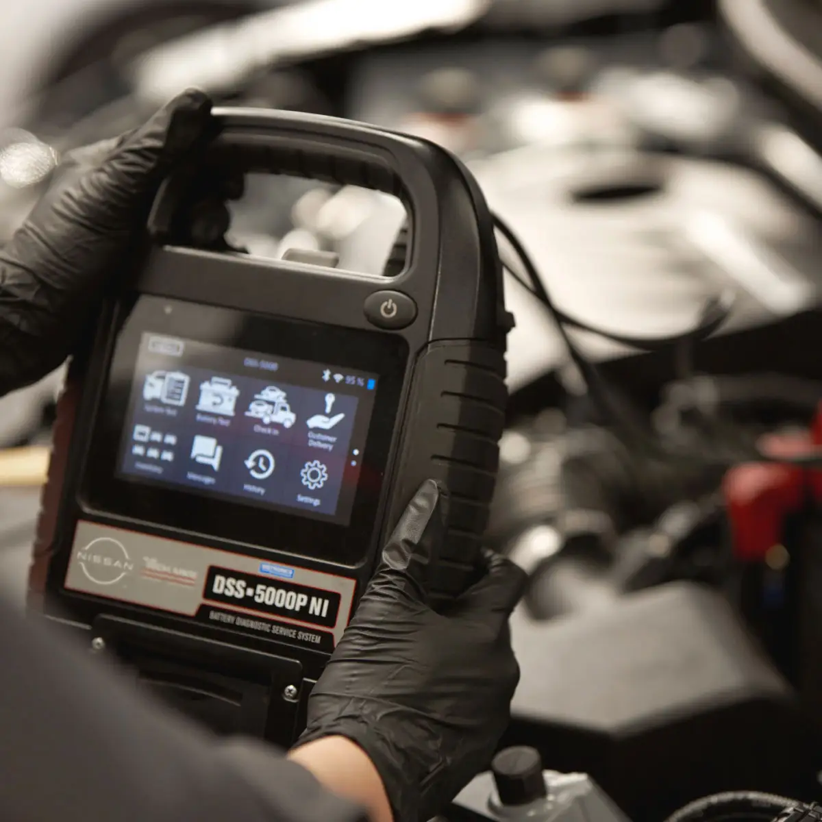 Nissan-trained technician holding portable car battery tester device connected to car battery