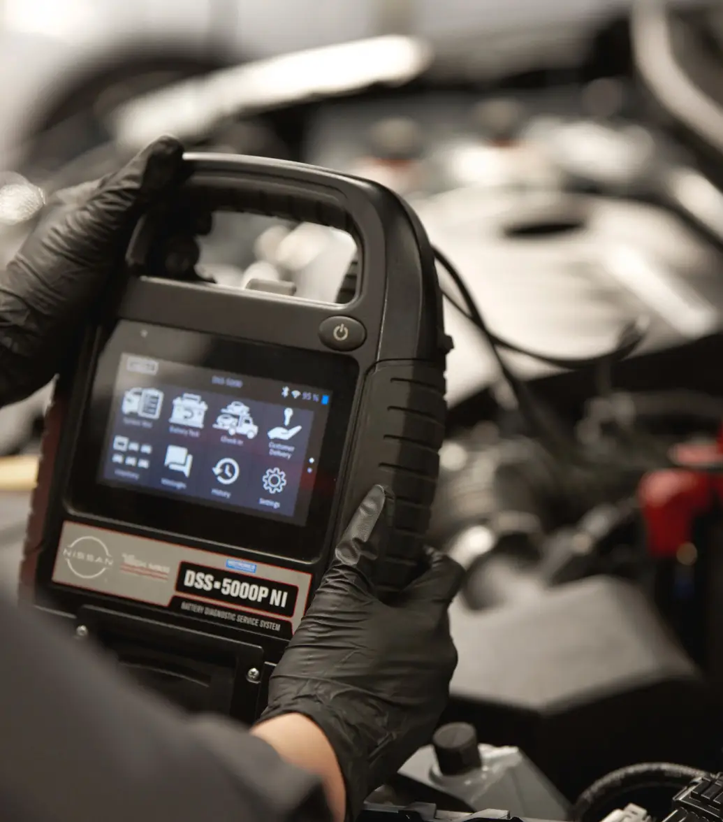 Nissan-trained technician holding portable car battery tester device connected to car battery