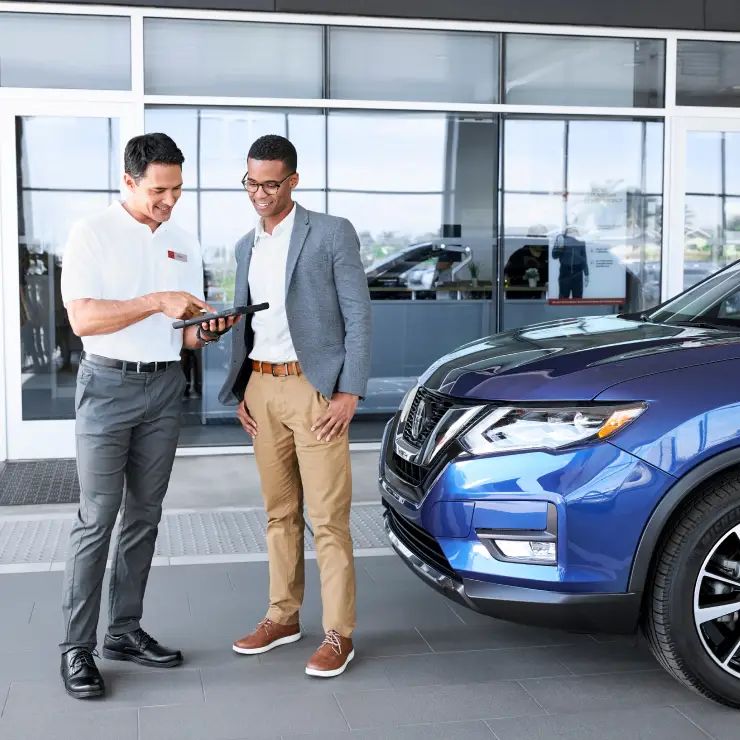 Salesperson showing a tablet to a smiling customer standing next to a blue SUV at a dealership.