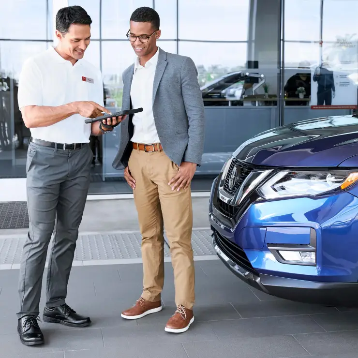 Two men standing next to a blue car, looking at a tablet and smiling.